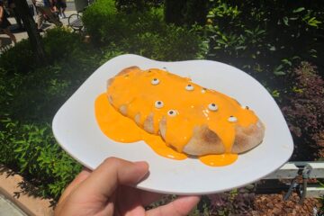 Plate with an oval Minion Cafe Cheesecake Calzone drenched in bright orange sauce and small white candy eyes, held outdoors near greenery.