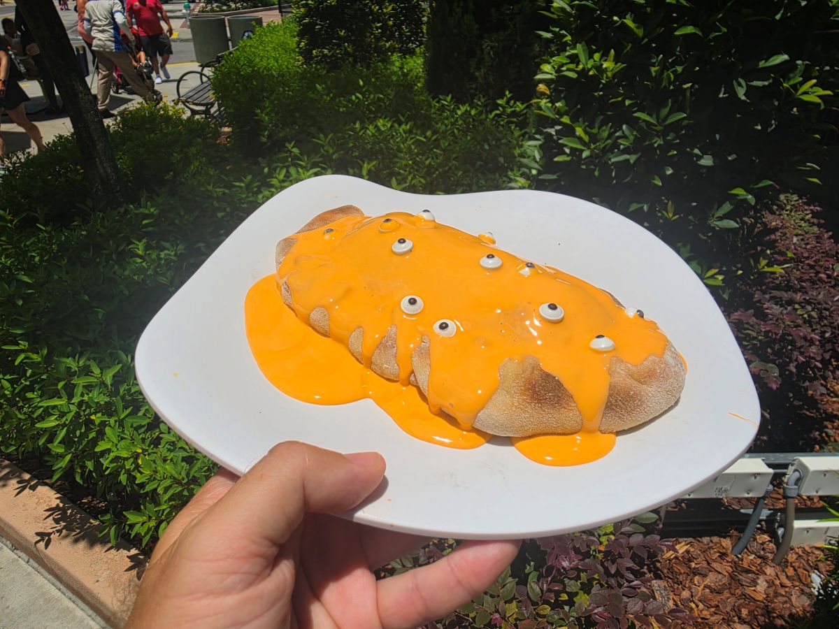 Plate with an oval Minion Cafe Cheesecake Calzone drenched in bright orange sauce and small white candy eyes, held outdoors near greenery.