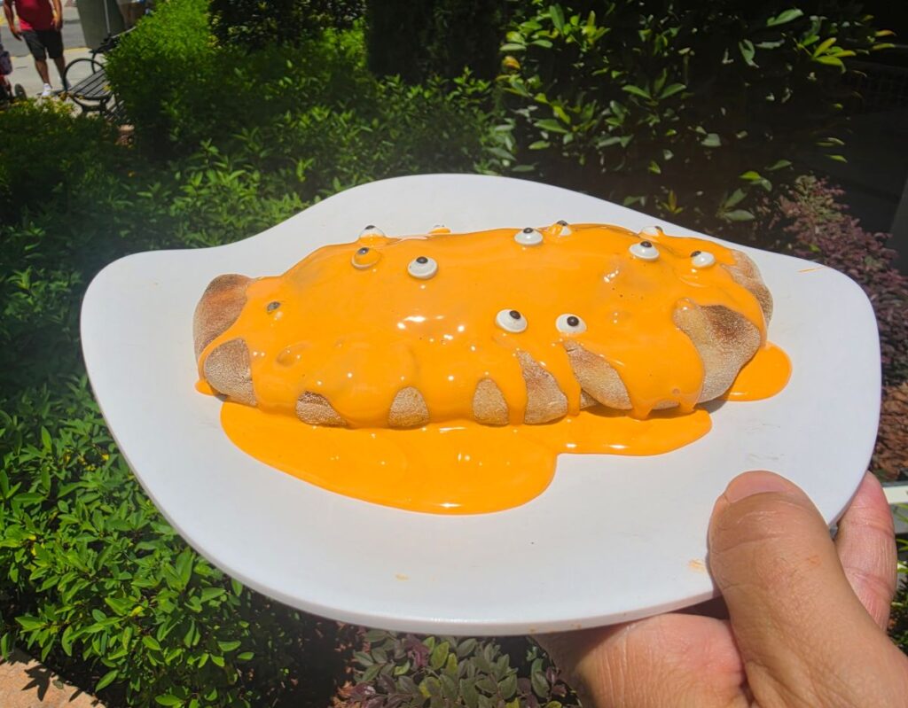 A hot dog-shaped Minion Cafe Cheesecake Calzone covered in orange cheese sauce with googly eyes on a white plate, held by a hand outdoors for a playful effect