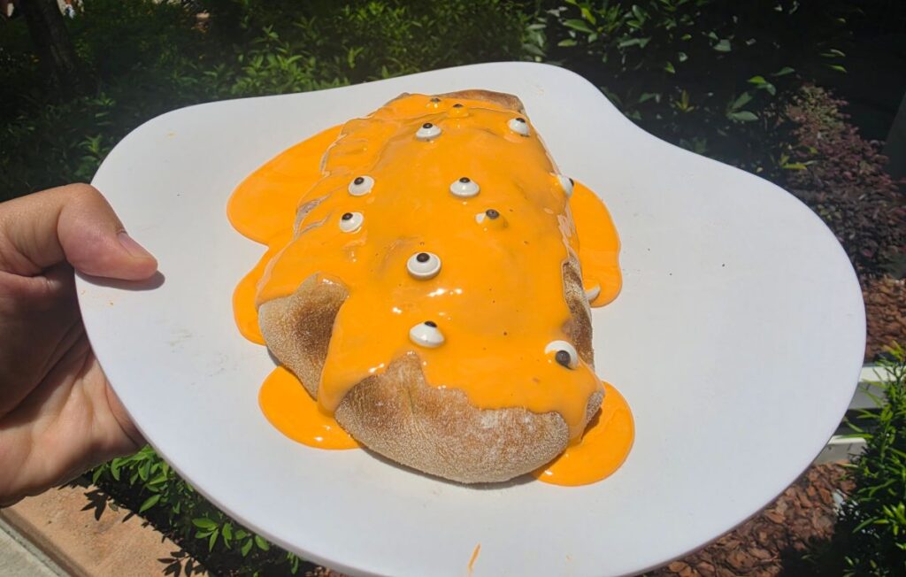 Orange-glazed Minion Cafe Cheesecake Calzone with candy eyes on a white plate outdoors, held by a hand.