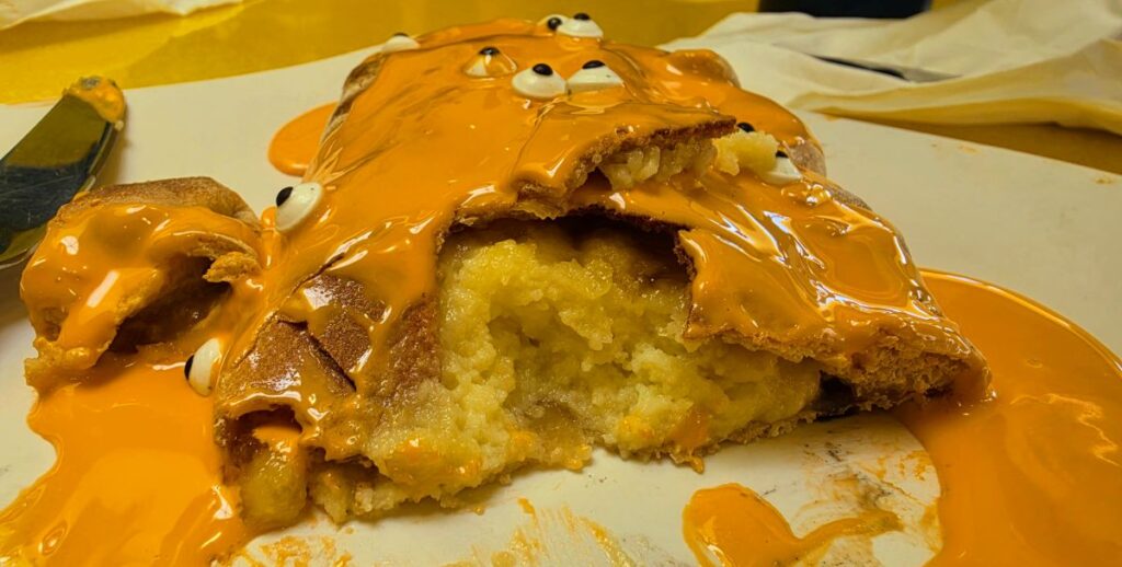 A sliced orange-frosted cake or pastry with gooey icing spilling over the plate and a bite taken out, exposing a pale yellow interior.