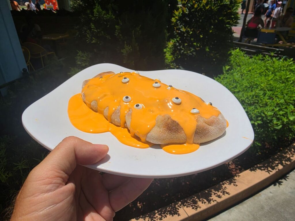 Hand holds a plate with a Minion Cafe Cheesecake Calzone covered in orange cheese sauce and candy eyeballs, outdoors in a sunny garden setting.