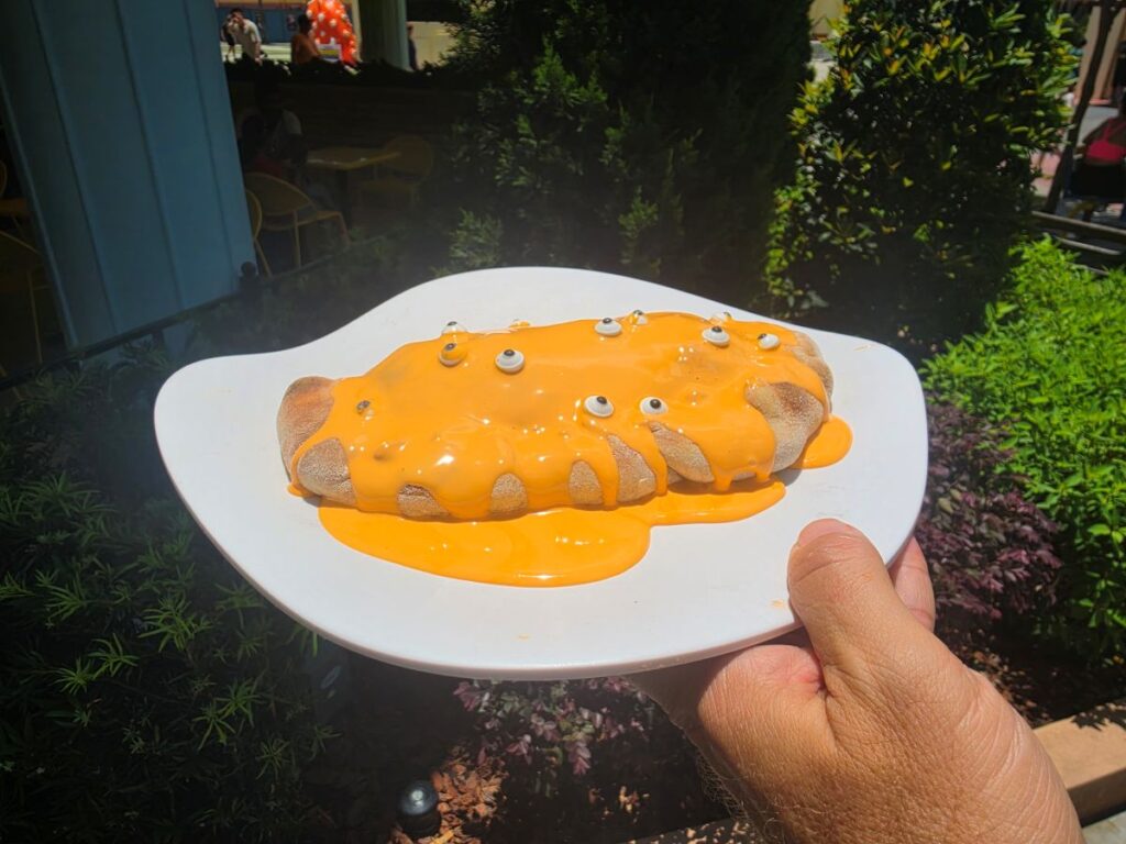 Plate held outdoors containing a Minion Cafe Cheesecake Calzone covered in orange cheese sauce with candy eyes on top.