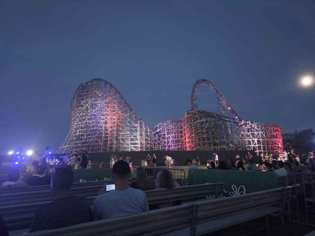 Nighttime view of a lit wooden roller coaster with purple and red lights, spectators seated on bleachers in the foreground.