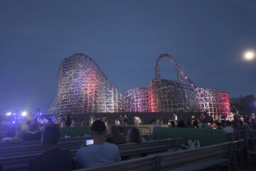 Nighttime view of a lit wooden roller coaster with purple and red lights, spectators seated on bleachers in the foreground.