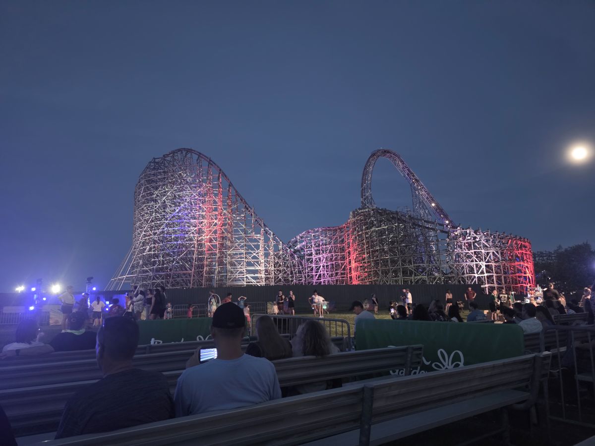 Nighttime view of a lit wooden roller coaster with purple and red lights, spectators seated on bleachers in the foreground.