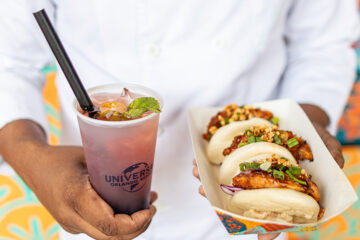 A person in a white shirt holds a pink gradient drink with ice and a black straw, featuring a Universal Orlando Resort logo, next to a tray of topped buns or bao filled with meat and veggies.