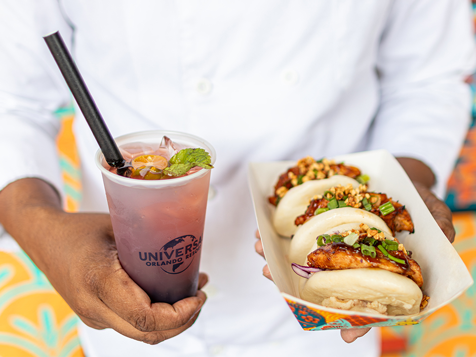 A person in a white shirt holds a pink gradient drink with ice and a black straw, featuring a Universal Orlando Resort logo, next to a tray of topped buns or bao filled with meat and veggies.