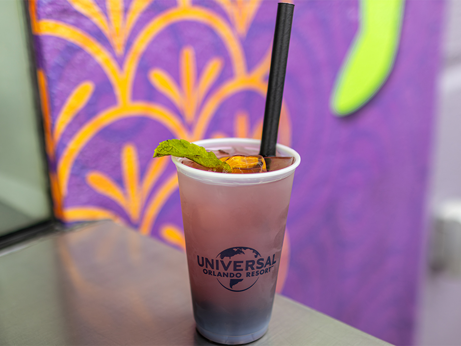 Cold pink-purple beverage in a clear cup with a black straw, mint garnish, and the Universal Orlando Resort logo visible on the cup, set against a colorful mural backdrop.