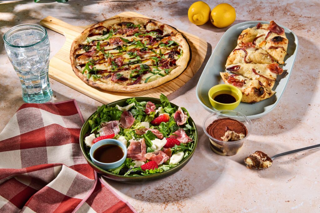Whole pizza with bacon and arugula on a wooden board, plus a plate of sliced pizza, a green salad, lemons, and a tall glass of water on a table promoting Italian Escape Menu at Blaze Pizza