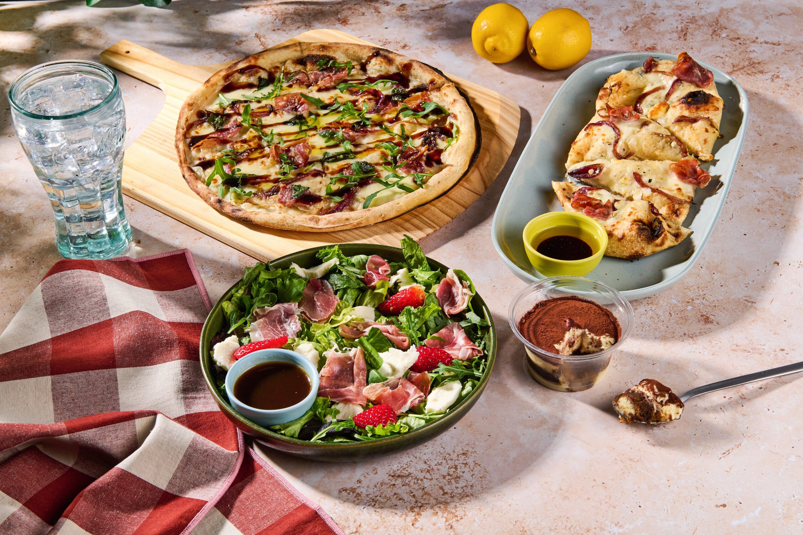 Whole pizza with bacon and arugula on a wooden board, plus a plate of sliced pizza, a green salad, lemons, and a tall glass of water on a table promoting Italian Escape Menu at Blaze Pizza