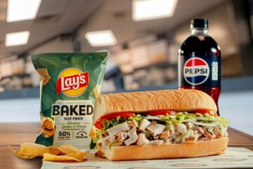 Jersey Mike's Chicken Salad Sandwich on a bun with chicken salad, lettuce, and tomato on a plate, with a bag of Lay's Baked Not Fried chips and a bottle of Pepsi in a casual dining setting.