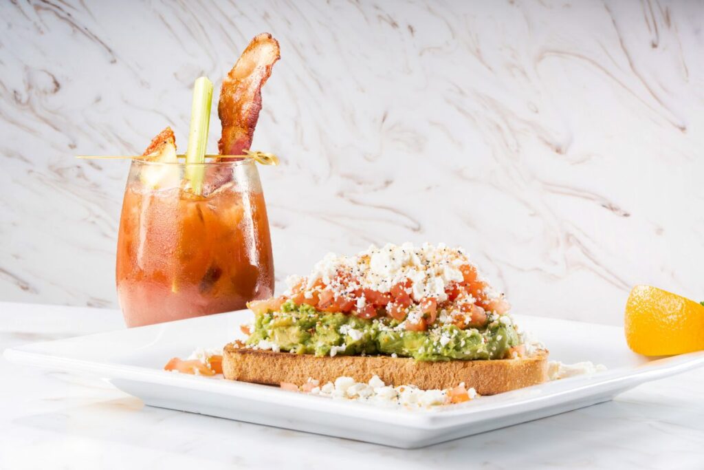 Brunch plate: avocado toast with tomato and feta on bread, served with a Bloody Mary garnished with celery and bacon to promote Keke's Summer Menu