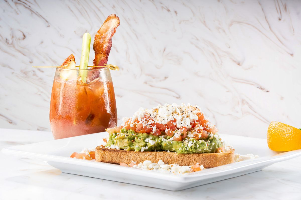 Brunch plate: avocado toast with tomato and feta on bread, served with a Bloody Mary garnished with celery and bacon to promote Keke's Summer Menu