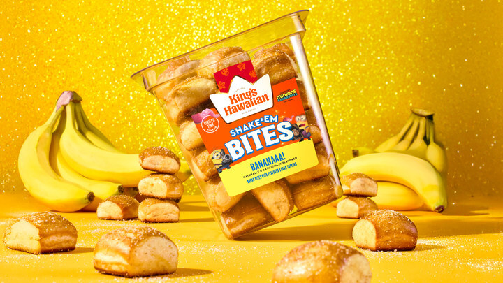 Tilted Kings Hawaiian Shake ’N Bites Banana flavor container on a bright yellow glitter backdrop, surrounded by bananas and bite-sized pastries to promote King's Hawaiian Banana Bites