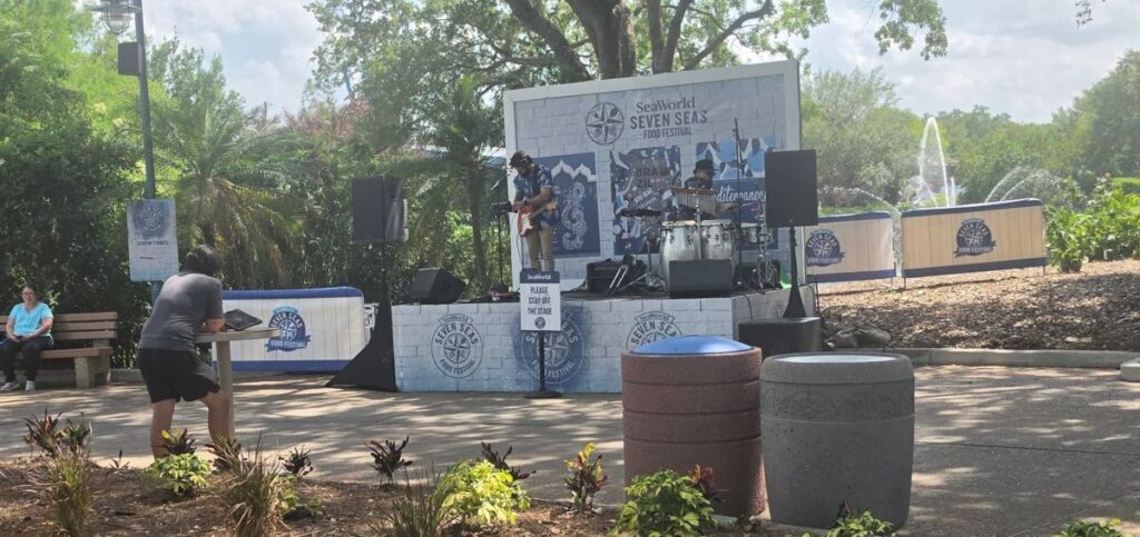 Outdoor festival stage with a band performing, SeaWorld Seven Seas Food Festival backdrop, trees in the background.