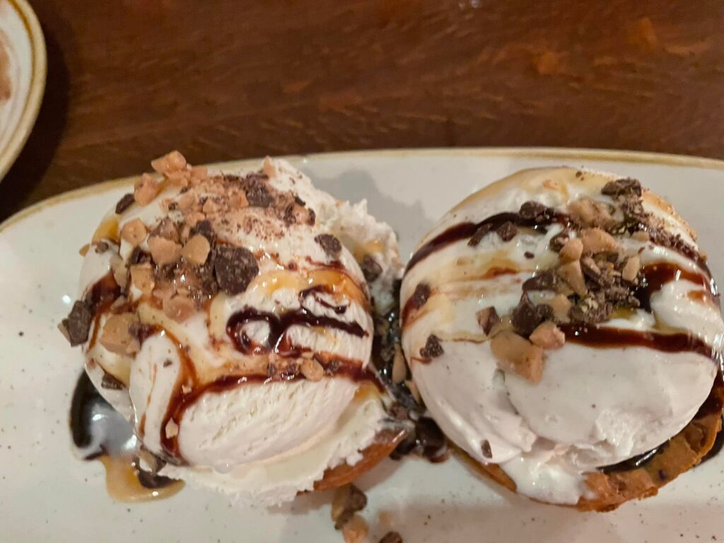 Two round pastries with white glaze, chocolate and caramel drizzles, and chopped nuts on a white plate.