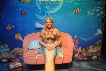 Mermaid-costumed woman sitting on a pink seashell couch in front of a Sea Life Orlando Aquarium backdrop with fish and coral imagery.