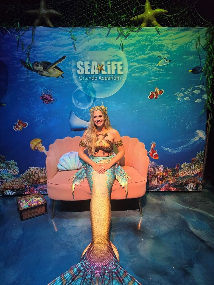 Mermaid-costumed woman sitting on a pink seashell couch in front of a Sea Life Orlando Aquarium backdrop with fish and coral imagery.