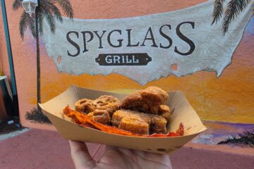Hand holding a disposable tray of Mickey Churro Waffles and bacon in front of a colorful mural that reads 'Spyglass Grill' with palm trees in the background.