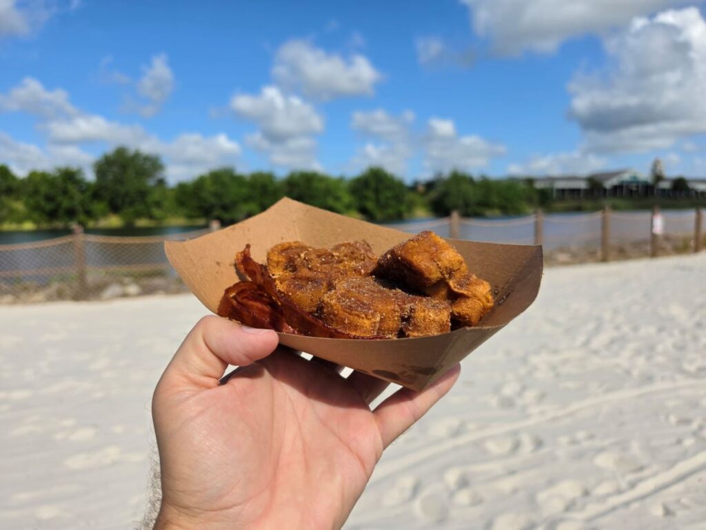Hand holding a brown paper boat filled with Mickey Churro Waffles and bacon on a sandy beach with a blue sky and a wooden fence in the background.