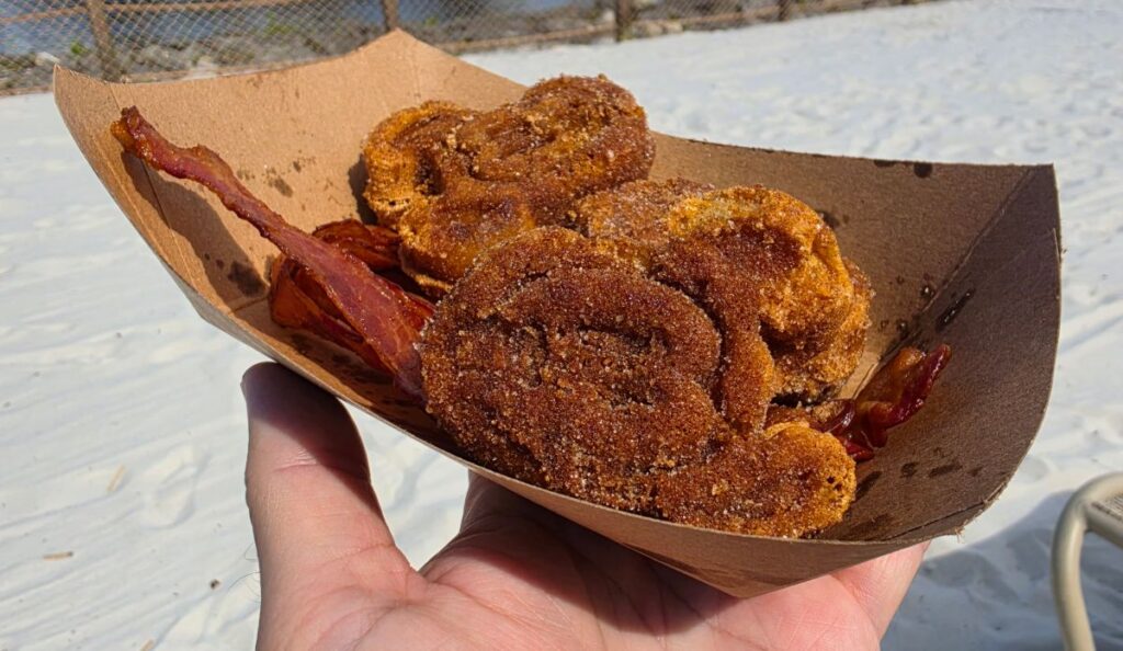 Hand holding a brown paper boat filled with Mickey Churro Waffles and a strip of crispy bacon, outdoors on sandy ground.