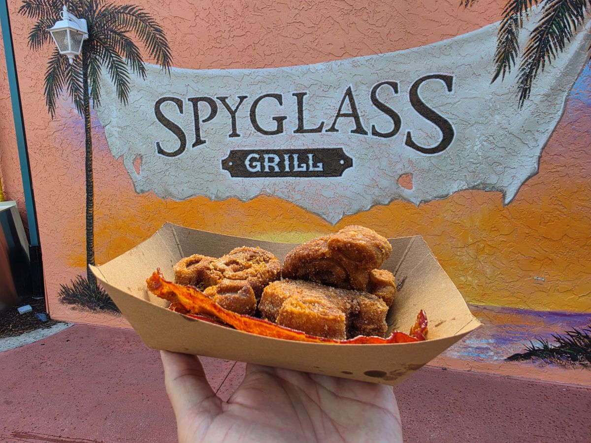 Hand holding a disposable tray of Mickey Churro Waffles and bacon in front of a colorful mural that reads 'Spyglass Grill' with palm trees in the background.