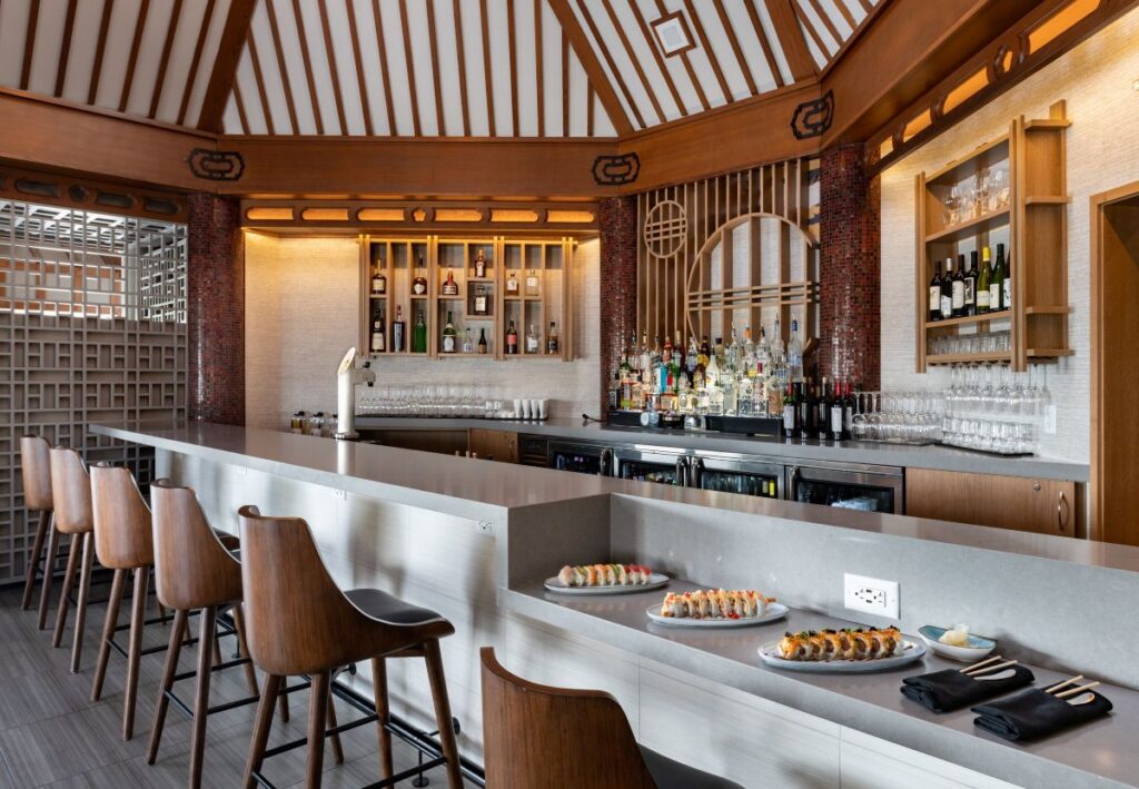 Modern sushi bar with a grey counter,wooden stools, and backlit bottles on display behind the counter; three sushi plates on the bar top. This place can be dined at using Orlando World Center Perks.