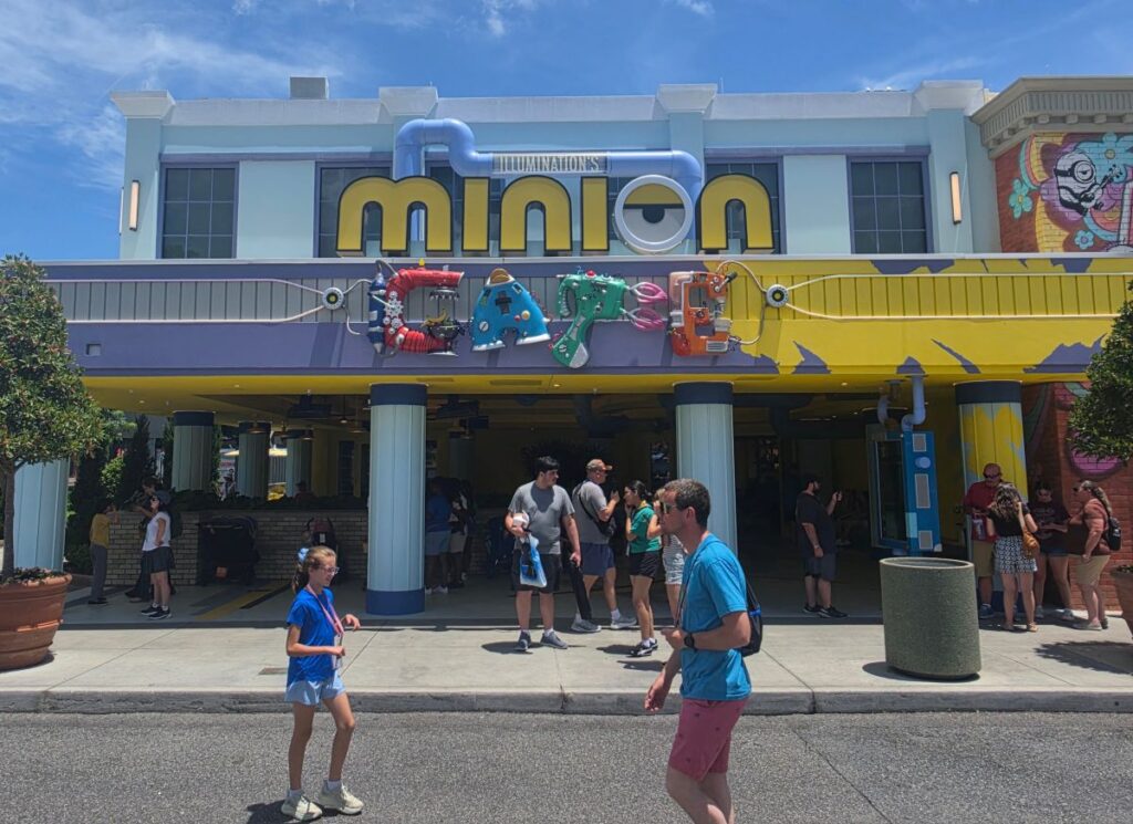 Front of a colorful amusement attraction with a large 'Minion' sign and playful tubing decorations, people walking outside on a sunny day.