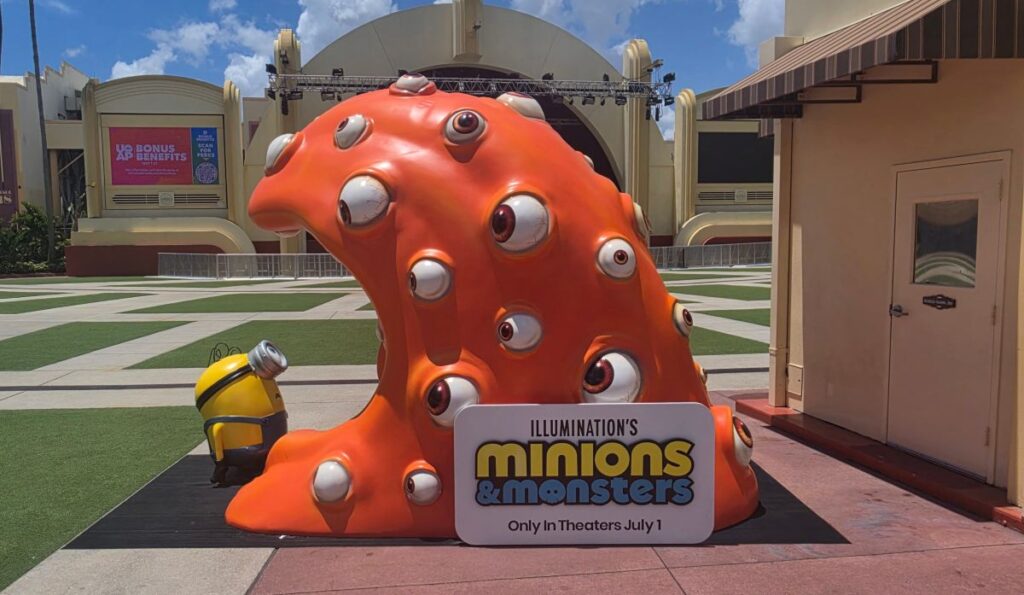 Large orange multi-eyed monster sculpture with a small Minion figure beside it, advertising Illumination's Minions & Monsters.