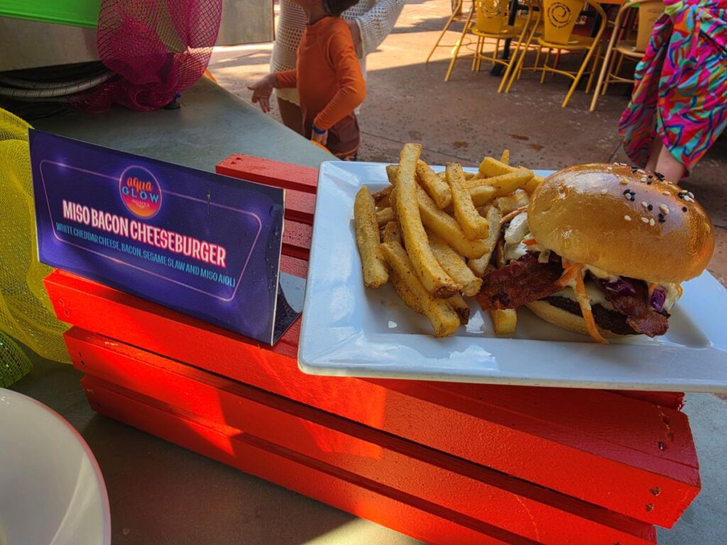 Plate with a miso bacon cheeseburger and fries on a white dish, resting on a bright red bench; sign reads 'Miso Bacon Cheeseburger' nearby.