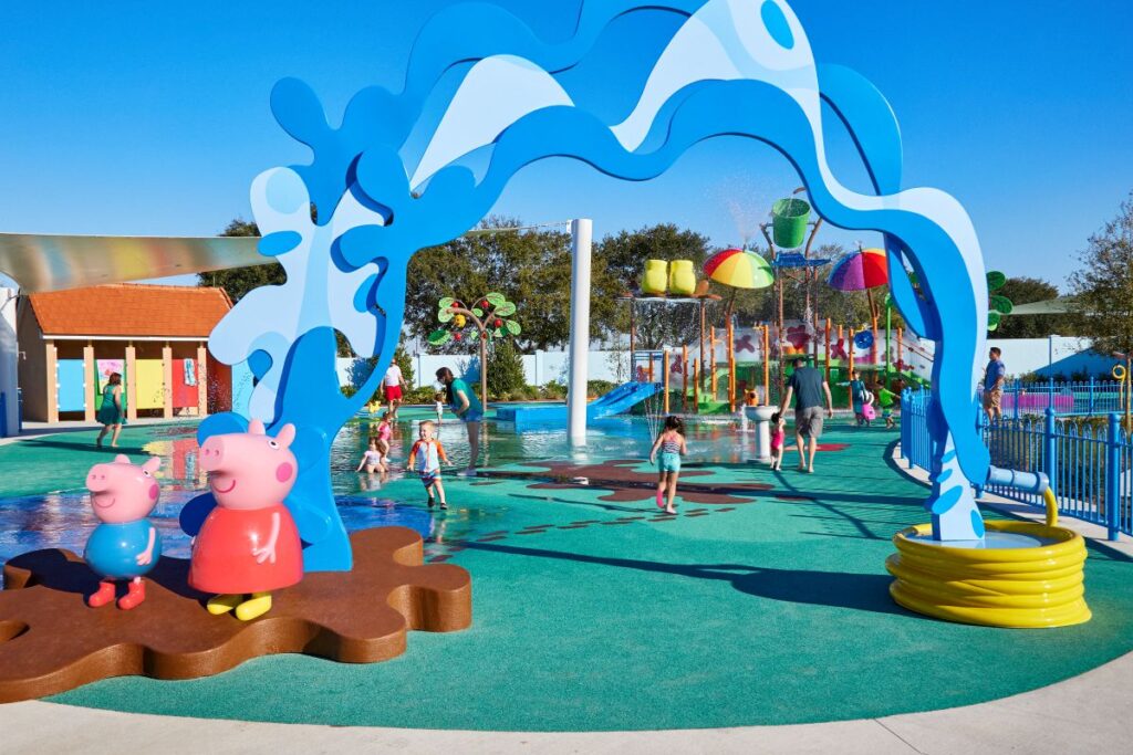 Colorful splash pad at a kids' water park with Peppa Pig statues on a brown stepping platform, a large blue abstract arch overhead, and children playing in shallow water beyond the sculptures.