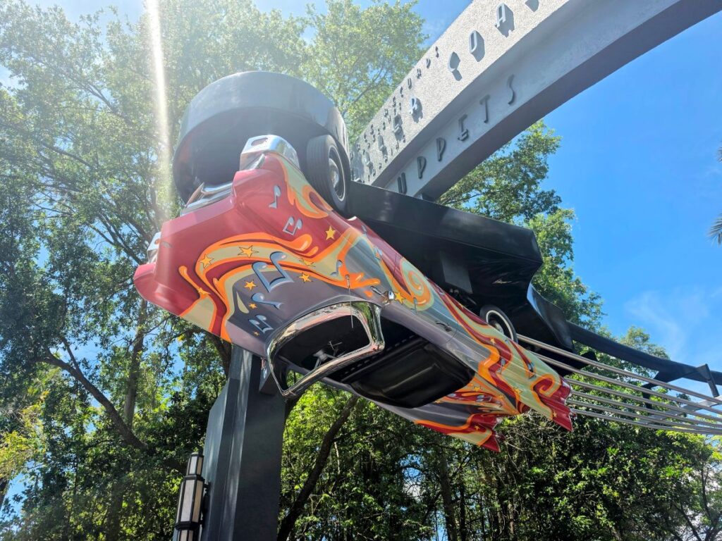 Upside-down colorful roller coaster car suspended on a tall arch, painted with orange and yellow flames against a blue sky and trees in the background for Muppets Rock 'n' Roller Coaster