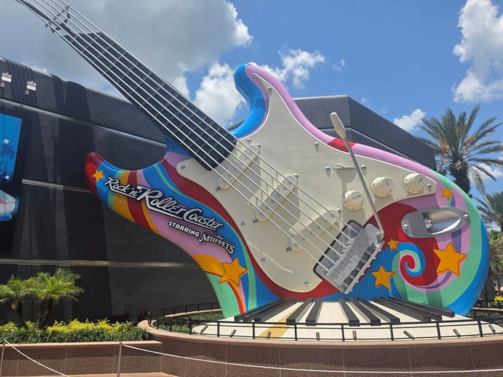 Giant colorful electric guitar sculpture reads 'Rock 'n' Roller Coaster' with 'Starring the Muppets', rainbow stripes and stars, outdoors with blue sky and palm trees in the background.