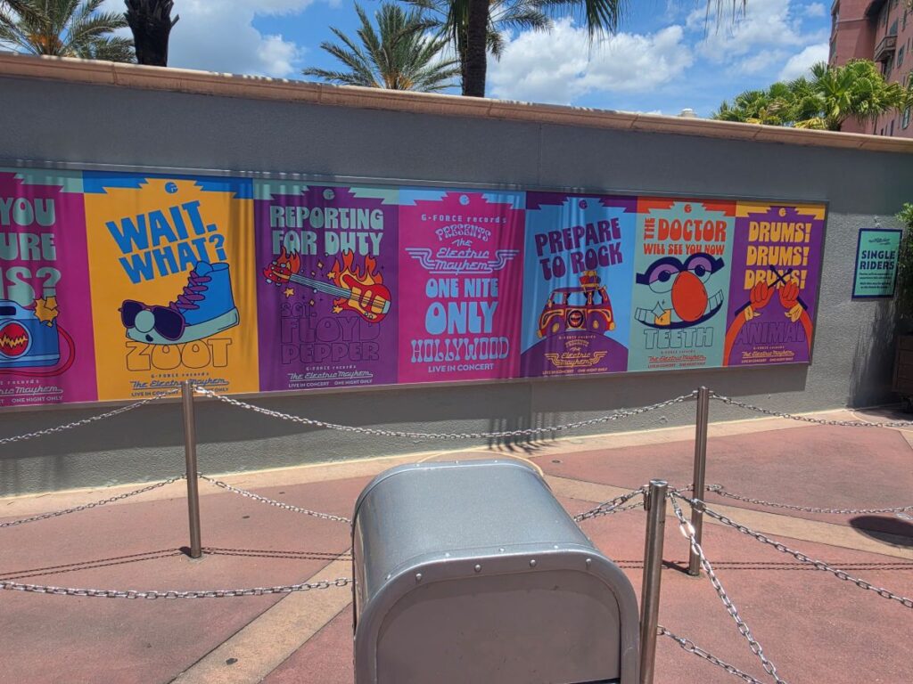 Row of bright, retro-style posters advertising concerts on a gray wall, with palm trees and blue sky in the background; a metal trash can and chain barriers are in the foreground.