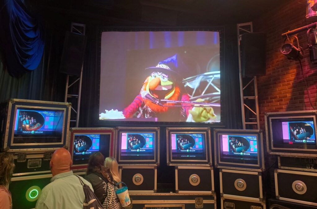 Stage setup with a large screen displaying a colorful muppet-style character performing, surrounded by smaller monitor desks and audience members watching nearby