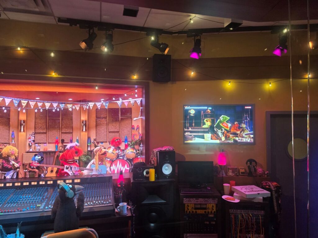 A colorful puppet band performing on a stage in a recording studio, with drums, guitars, and bunting hanging above.