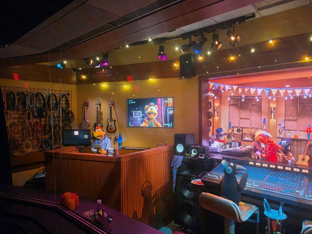 Muppet show recording studio: two puppets at a wooden control desk, with guitars on the wall, a monitor, and a large mixing console under colorful lights