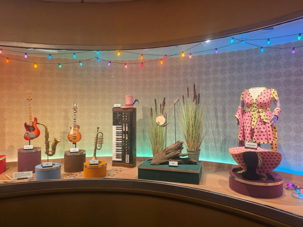 Display case with musical instruments (guitars, saxophone, keyboard) and a pink polka-dot dress on a mannequin, plus a matching hat, under colorful string lights in queue for Muppets Rock 'n' Roller Coaster