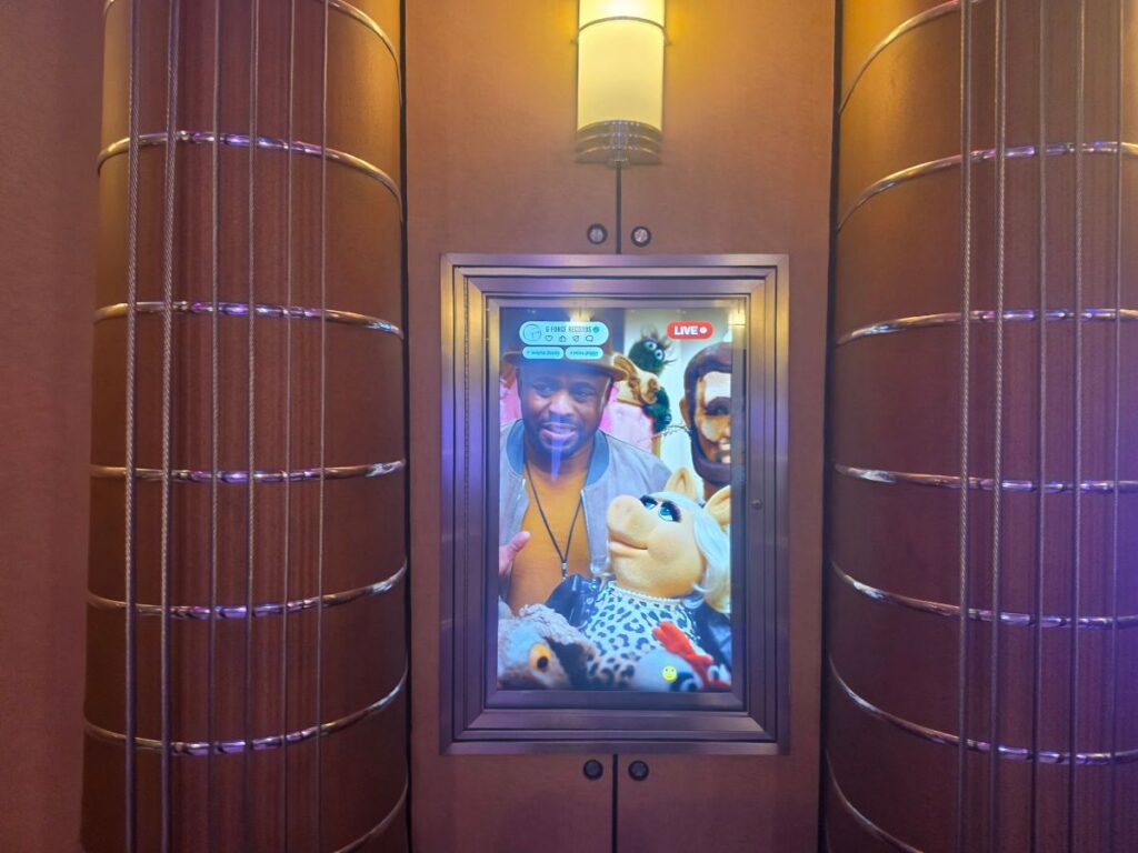 Digital display in an elevator showing a man in a yellow shirt and blue jacket with colorful stuffed animals around him; a LIVE badge visible on the screen.