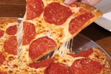 Hand lifting a pepperoni pizza slice of Marco's NY Style Pizza with melted cheese stringing from the slice on a wooden table edge.