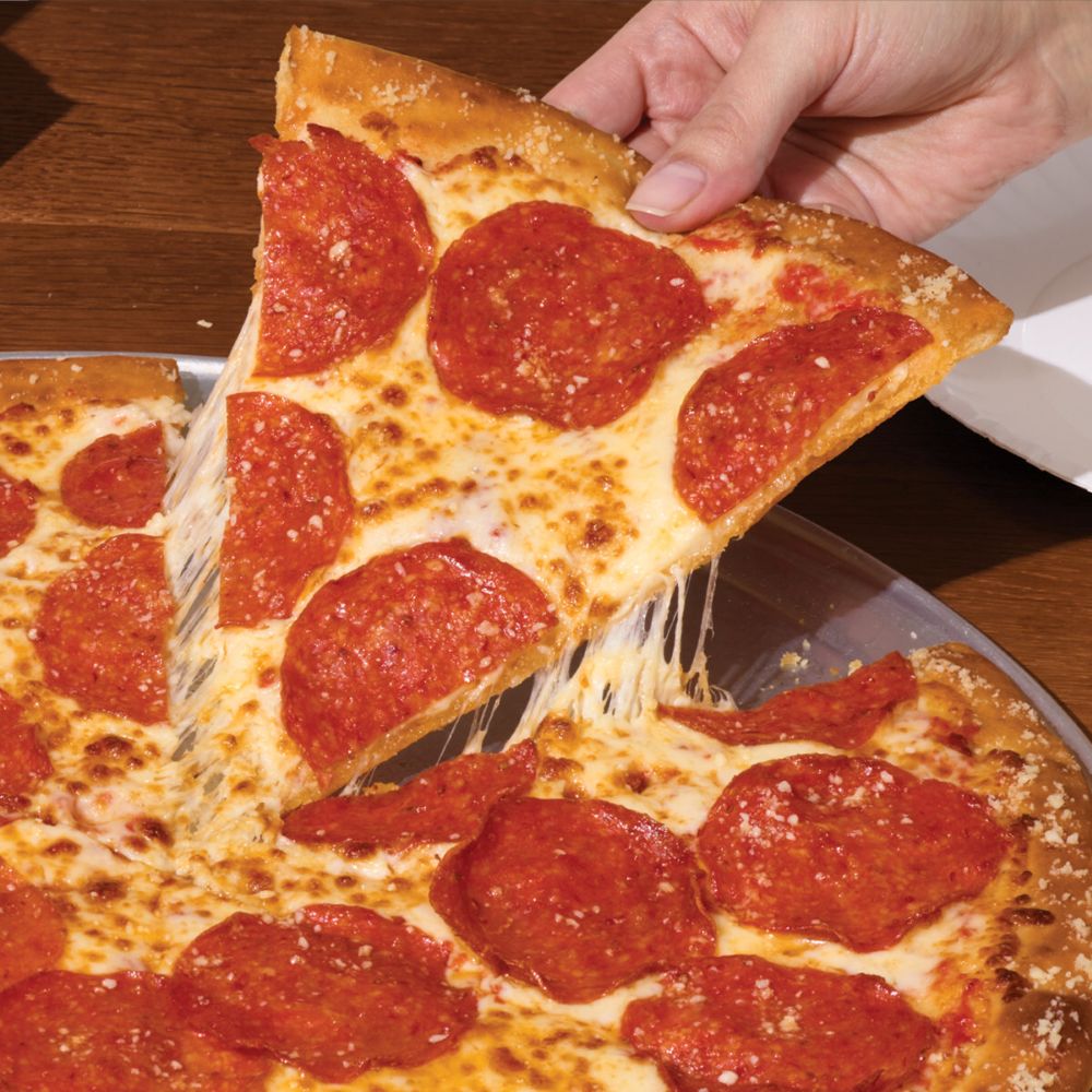 Hand lifting a pepperoni pizza slice of Marco's NY Style Pizza with melted cheese stringing from the slice on a wooden table edge.
