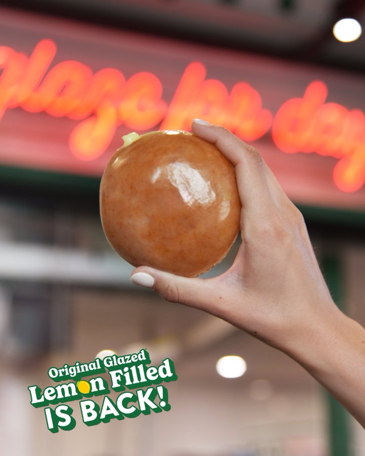 Close-up of a hand holding a glossy lemon-filled original glazed donut with a blurred neon sign in the background and promo text 'IS BACK'.