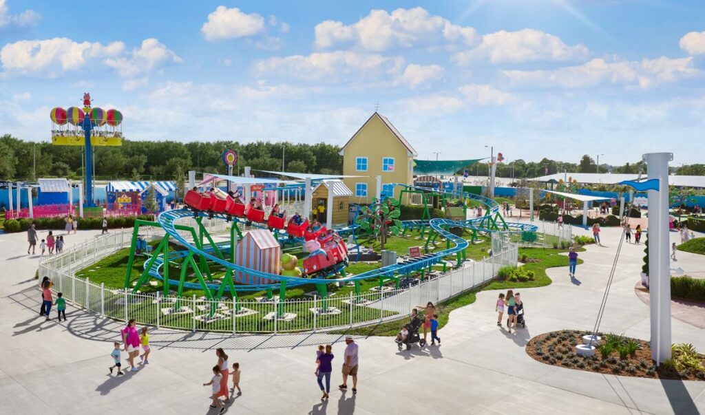Colorful amusement park with a looping roller coaster surrounded by a white fence, sunny sky, and visitors strolling on the paved paths.