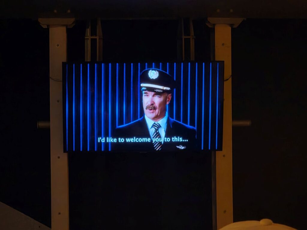 TV screen showing a man in a captain uniform speaking, with the subtitle 'I'd like to welcome you to this...'.