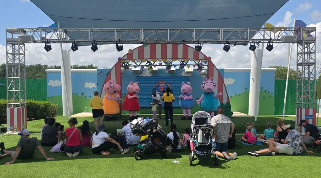 Outdoor stage show with five colorful mascots entertaining a seated crowd on a grassy lawn under a canopy at Peppa Pig Theme Park