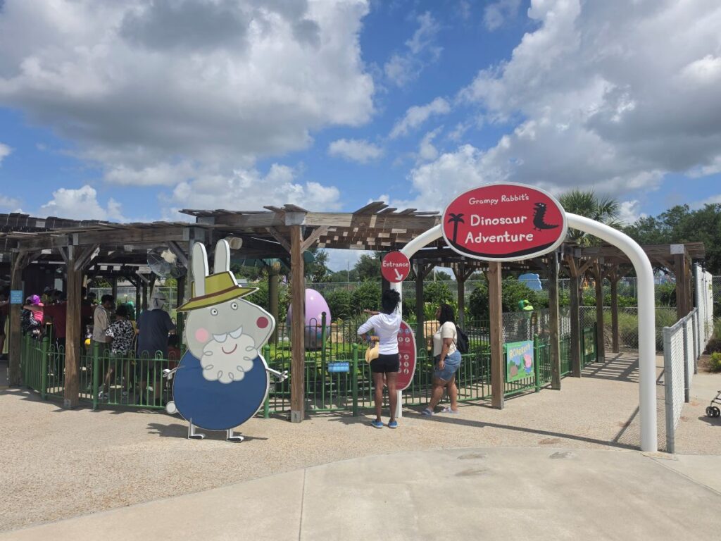 Entrance to Grumpy Rabbit's Dinosaur Adventure ride, with a large cartoon rabbit cutout at the front and people queuing under wooden shade structures.