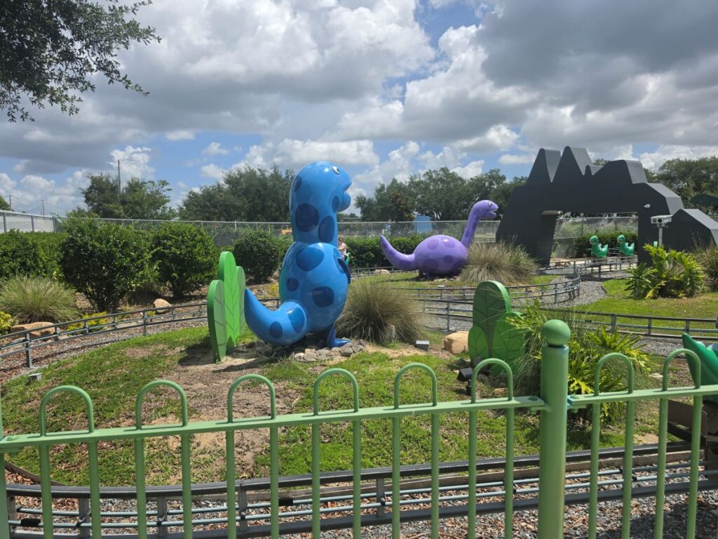 Colorful dinosaur sculptures along a miniature train track in a fenced outdoor play area, with blue and purple dinos and a tunnel in the background.
