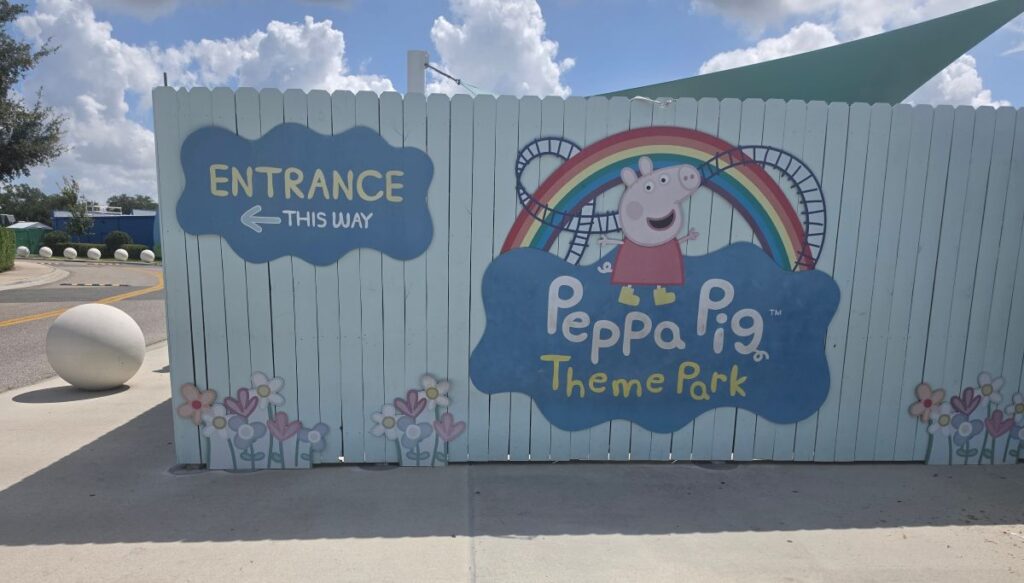 Entrance sign for Peppa Pig Theme Park featuring a rainbow and Peppa Pig illustration on a light blue fence with flowers.