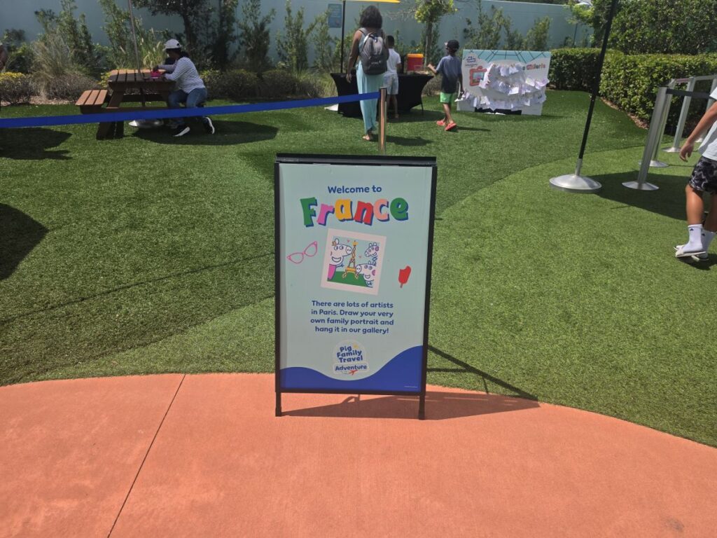 Outdoor event scene with a vertical sign reading 'Welcome to France' on a grassy area, people chatting in the background near tables and a fence.
